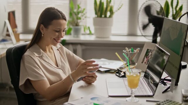Medium shot of Caucasian woman sitting at desk, drinking orange juice slowly while texting on social network, breaking into smile during workday in office