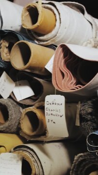 Vertical video. Rolled fabric and upholstery materials stacked in textile workshop storage. Close-up shot of textured fabric rolls, labels, layered textiles and sewing production supplies. Filmed in
