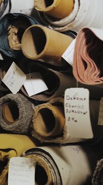 Vertical video. Textile material rolls and upholstery fabric stored in workshop shelves. Close-up shot of layered fabric textures, rolled cloth, paper labels and sewing industry supplies. Filmed in 4K