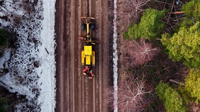Motor grader leveling dirt road in a forest between winter and spring in aerial top down tracking shot