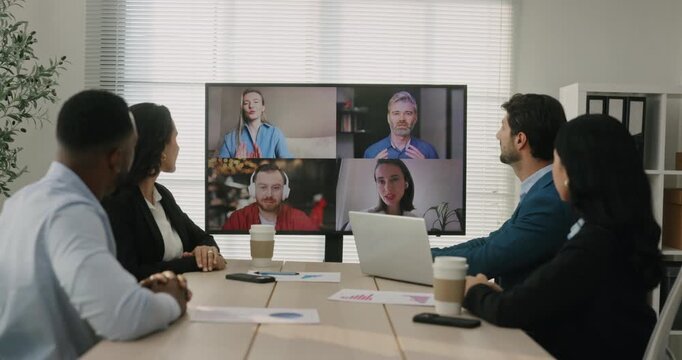 Multicultural office team joins video meeting with remote coworkers on large screen in boardroom. Colleagues watch display and discuss project updates together