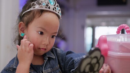 A young toddler girl wearing a tiara plays with makeup, exploring creativity and self-expression during her childhood development in a close-up portrait. © HarryKiiM Stock