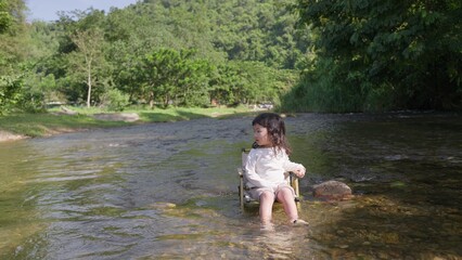 Fototapeta premium A young child sits peacefully on a camping chair in a shallow river, enjoying a quiet moment of sensory play and connecting with the natural environment.