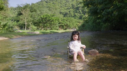 Fototapeta premium A young child sits peacefully on a camping chair in a shallow river, enjoying a quiet moment of sensory play and connecting with the natural environment.