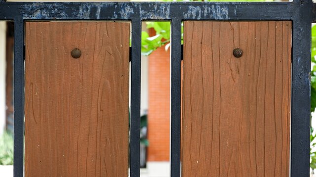 Close-up architectural shot of a modern residential fence gate, featuring vertical brown wood grain textured planks mounted securely within a weathered black iron metal frame profile.
