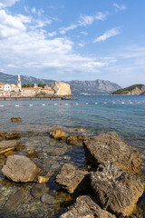 Obraz premium Sunny coastal view of Budva old old town with stone fortress, red-tiled roofs and bell tower overlooking clear Adriatic waters, rocky shoreline and distant mountains