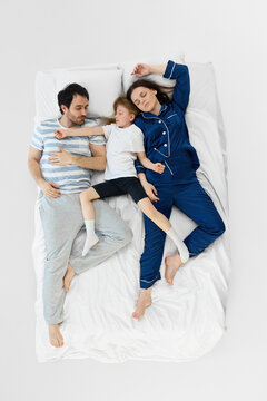 Overhead view of family sleeping together on white bed with calm home lifestyle mood. Concept of healthy sleep, parenting, family bonding, bedroom comfort, and wellness advertising.