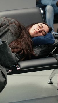 Vertical video Tired woman in airport lounge sleeping on metal bench during long airplane layover. Jet lagged traveler resting on seats, napping on neck pillow while waiting for delayed plane, camera