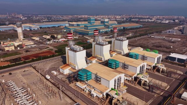 Aerial view of a combined cycle gas power plant featuring gas turbines, heat recovery steam generators, exhaust stacks emitting steam, and a high-voltage electrical switchyard in an industrial zone