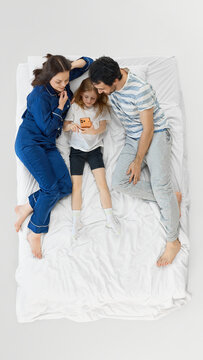 Happy family relaxing in bed while daughter uses smartphone between smiling parents. Concept of family screen time, bedding ads, mattress comfort, digital parenting, and weekend home lifestyle.