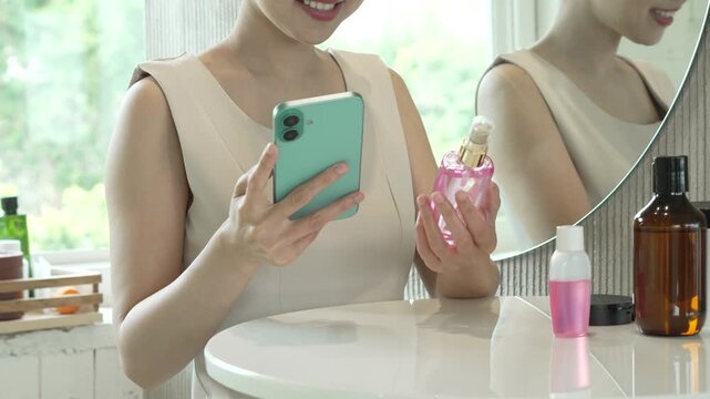 Smiling woman at vanity holding smartphone and pink perfume bottle beside cosmetics in bright bathroom, reflected in mirror, modern lifestyle beauty routine.