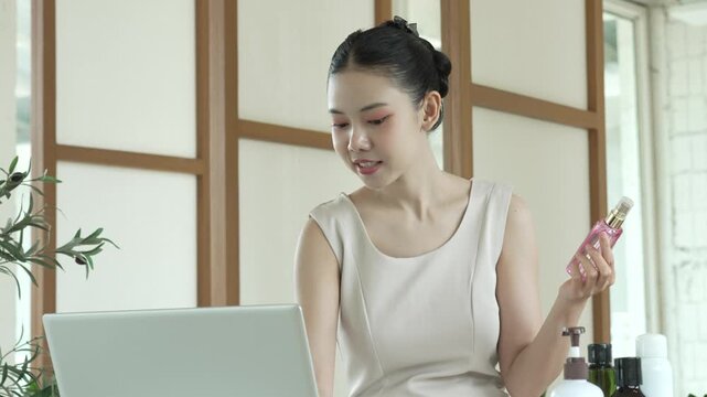 Young woman at home using laptop while holding perfume bottle, skincare products on table, bright minimalist interior, self-care and online shopping concept.