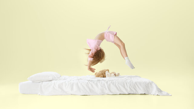 Girl doing backflip above white bed with teddy bears in playful studio bedroom scene. Concept of active childhood, playful bedroom lifestyle, mattress comfort, and kids bedding ads.