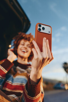 Smiling woman lifestyle outdoors, holding smartphone for a selfie, warm sunset light, soft focus background with sky and road, casual striped sweater, playful moment near vehicle, natural colors