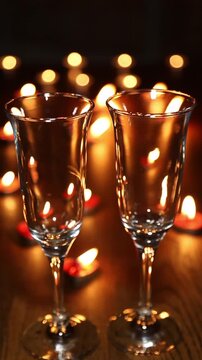 Two empty crystal wine glasses stand on a wooden table surrounded by warm glowing bokeh candles. featuring elegant glass reflections and warm amber tones.
