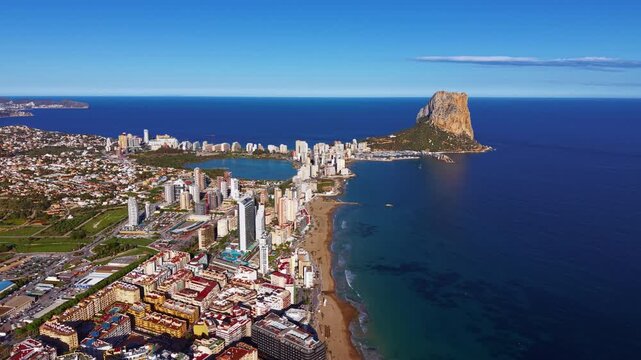Aerial view of Calpe, Spain showing the iconic Penon de Ifach limestone rock, sandy beach, coastal city skyline, salt lake lagoon, and deep blue Mediterranean Sea