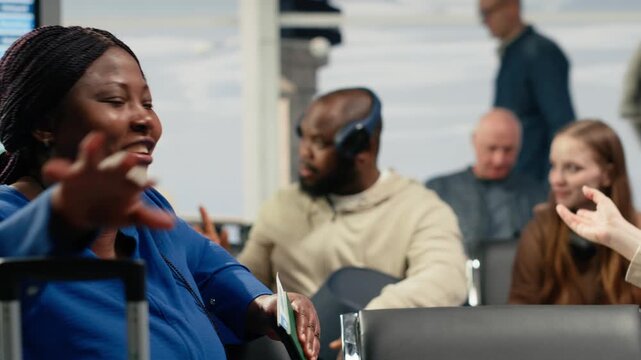 Travel friends sit in an airport waiting area and talk with expressive gestures while other passengers wait behind them near runway windows.