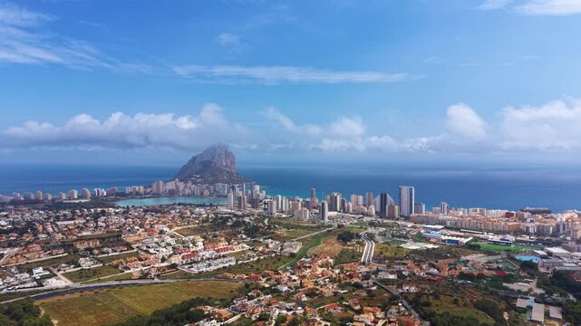 Panoramic elevated view of Calpe, Spain, featuring the iconic Penon de Ifach limestone rock, coastal skyline, residential areas, and blue Mediterranean Sea under partly cloudy sky