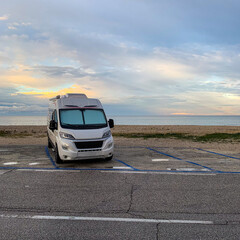 A white modern recreational vehicle camper van parked in a designated parking lot next to a pebble beach, looking out over a calm sea under a beautiful sunset sky. © Licia.Galli
