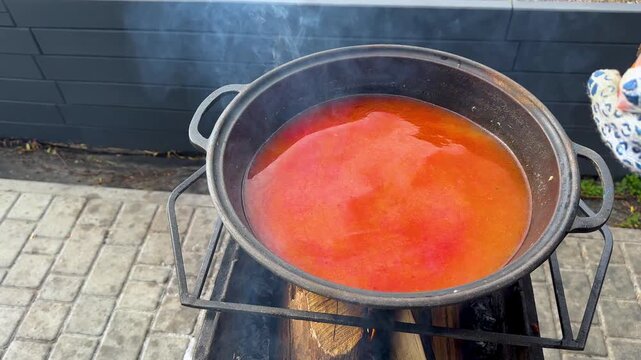 Stirring Tom Yum paste into chicken broth in a large cast iron cauldron over an open wood fire outdoors