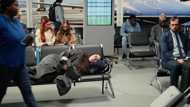 A tired woman sleeps across metal seats in an airport lounge during a long layover or flight delay while other travelers pass through the terminal.