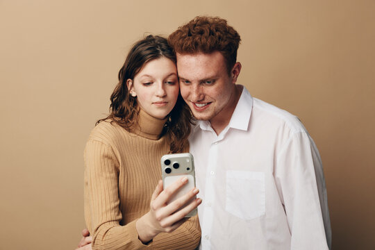 Young couple with curly hair takes selfie together using smartphone on beige background. They smile and enjoy moment, showing affection and modern lifestyle with technology.