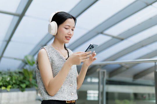 A young woman wearing headphones uses her smartphone indoors, appearing focused and engaged in a modern setting.
