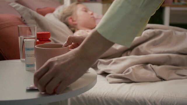 Unknown woman bringing mug of hot tea and blister pack, placing them on side table, touching forehead of coughing daughter and adjusting her blanket