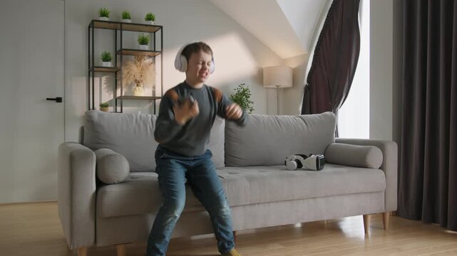 Young boy wearing headphones listens to music on his phone while sitting on a comfortable sofa in a modern living room. The calm moment conveys childhood relaxation, digital enjoyment and the warmth o