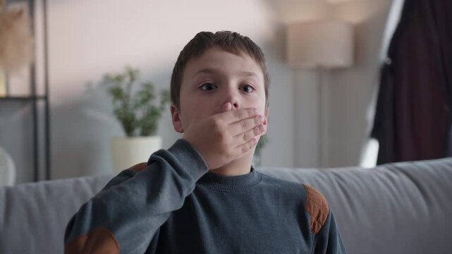 Young boy with wide eyes covers his mouth with his hand, expressing surprise or shock while sitting on a couch. The expressive moment highlights childhood innocence, emotion and the spontaneity