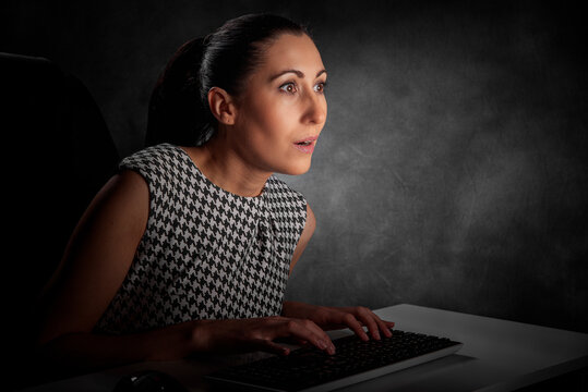 A surprised businesswoman sits at her desk at night, staring in shock at her glowing computer screen.