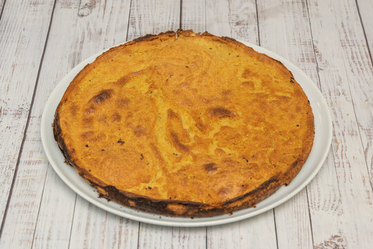 A round thin faina chickpea flatbread with golden bubbly surface sits centered on a white plate over whitewashed wooden planks.