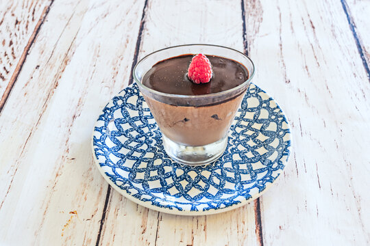 Creamy chocolate mousse topped with a fresh raspberry sits in a glass cup on a decorative blue and white plate.
