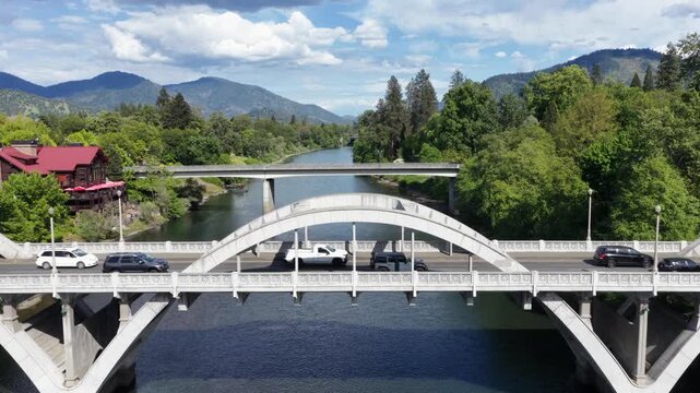 Grants Pass Oregon white concrete arch bridge crossing calm Rogue River with traffic, red cabin restaurant, lush green trees and blue mountain ranges under sunny blue sky. Aerial rising wide shot.
