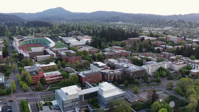 University of Oregon campus in Eugene with Autzen Stadium, red brick academic buildings, lush green trees and rolling mountain ranges stretching under soft overcast morning sky. Aerial rise pull back