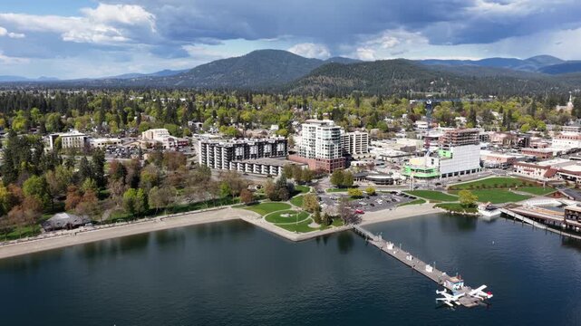 Coeur d'Alene lakefront with sandy beach, green parks, modern residential high-rises, charming downtown and dense pine forest stretching toward blue mountain ranges. Aerial lateral wide shot.