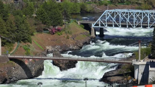 Spokane River Falls in Washington State with powerful rushing water and white splashes flowing under historic city bridges and pedestrian walkways during sunny day in USA. Aerial wide shot.