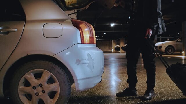 A lone figure with long hair loads a suitcase into the trunk of his car in a dark, quiet parking garage.