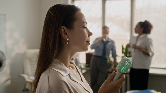 Brunette woman switching on portable mini fan, breaking into smile while enjoying wind while her colleagues communicating in blurred background