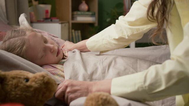 Caring long-haired mother pulling up daughters blanket and giving her kiss while sick girl napping in bed