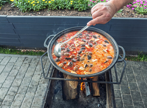 Cooking spicy Tom Yum soup in a large cast iron kazan over an open wood fire outdoors, hand stirring with a ladle