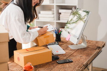 Small Business Owner Working: A dedicated female entrepreneur diligently organizes packages and writes on a clipboard, surrounded by her workspace filled with shipping boxes and a computer. © Kritdanai