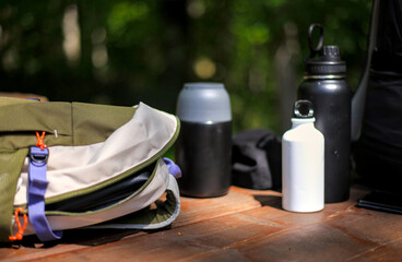 Close-up of a hiking backpack on a wooden table with reusable water bottles and a blurry green forest background. Great for outdoor, travel, and active lifestyle themes. © YuliiaKettu