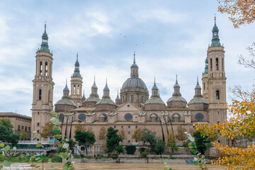 Fototapeta premium Birds fly over the magnificent towers of the Basilica of Our Lady of the Pillar in Zaragoza. Close-up view features colorful tiled domes and autumn branches. Zaragoza, Spain - November 29, 2021