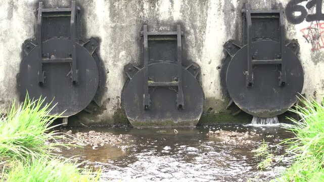 Water Management Infrastructure: Drainage Sluice Gates and Culvert Flow, Krakow, 2026, spring.The camera cuts to a tight, steady close-up shot of the central, slightly smaller metal flap gate