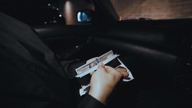 Anonymous criminal man sitting in a car counting a stack of hundred dollar bills from an envelope