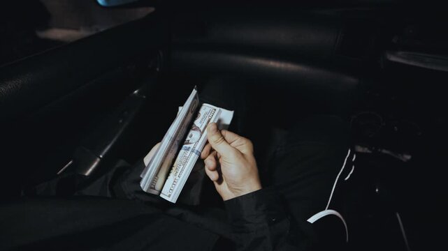 An anonymous first-person shot shows a man checking an envelope containing a large stack of hundred-dollar bills inside a car.