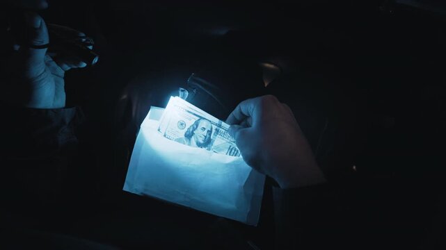 Hands of a man checking and recounting hundred dollar bills inside an envelope in a dark car