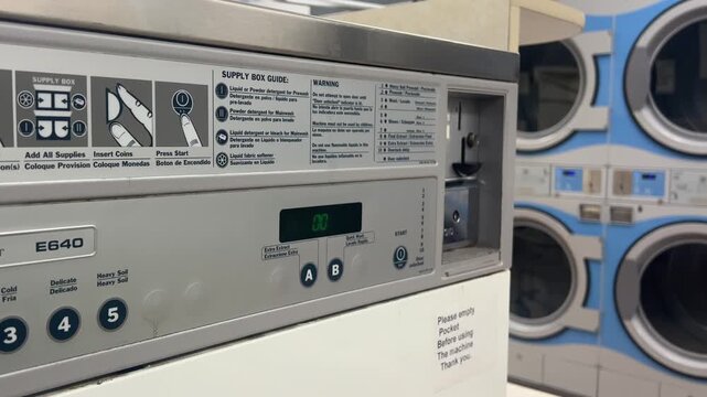 A close-up of a washing machine's control panel showing wash options and a coin slot in a laundromat during the day