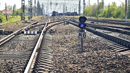 Naklejka premium Railway tracks curving through a rural area. A blue signal light is visible beside the tracks. The scene is set in a sunny environment with greenery.